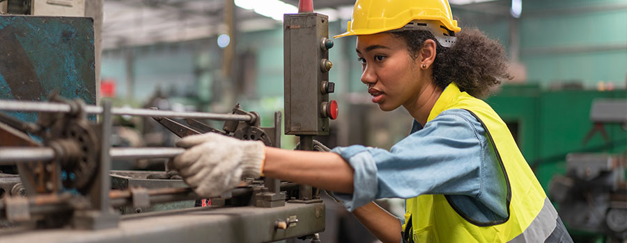 black woman wearing a hard had and reflective vest working a factory machine